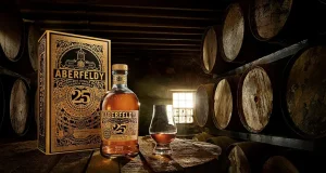 Aberfeldy® Celebrates 125 Years Of Crafting The Finest Scotch Whisky With Release Of A 25 Year Old Expression a bottle of Aberfeldy 25-year-old single malt scotch whisky beside its ornate box, prominently displayed on a wooden barrel in a dimly lit barrel room. To the right of the bottle, there's a whisky glass, filled with amber liquid, capturing the essence of the aged spirit. Rows of aged whisky barrels are stacked in the background, their curved shapes receding into the shadows, adding depth and a sense of tradition to the scene. A small window with a grid lets in a stream of light, illuminating the dust in the air and highlighting the product.