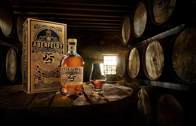 a bottle of Aberfeldy 25-year-old single malt scotch whisky beside its ornate box, prominently displayed on a wooden barrel in a dimly lit barrel room. To the right of the bottle, there's a whisky glass, filled with amber liquid, capturing the essence of the aged spirit. Rows of aged whisky barrels are stacked in the background, their curved shapes receding into the shadows, adding depth and a sense of tradition to the scene. A small window with a grid lets in a stream of light, illuminating the dust in the air and highlighting the product.
