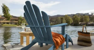 Deep blue: BeautiTone unveils 2024 Exterior Colour of the Year, ‘Pacific’ A deep blue Adirondack chair with a bright orange throw blanket draped over one arm rests on a wooden dock. Beside the chair is a small round wooden table holding a wine glass and a vintage-looking radio. In the background, a serene lake with hills and houses under a clear sky creates a peaceful setting. A striped tote bag stands next to the chair, suggesting a leisurely day by the water.