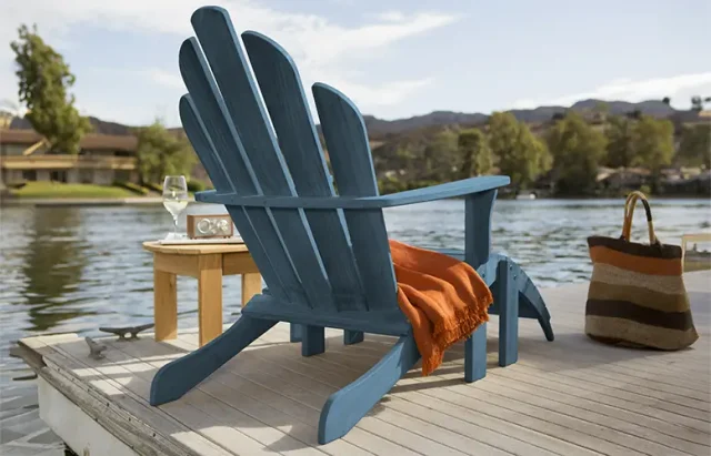 A deep blue Adirondack chair with a bright orange throw blanket draped over one arm rests on a wooden dock. Beside the chair is a small round wooden table holding a wine glass and a vintage-looking radio. In the background, a serene lake with hills and houses under a clear sky creates a peaceful setting. A striped tote bag stands next to the chair, suggesting a leisurely day by the water.